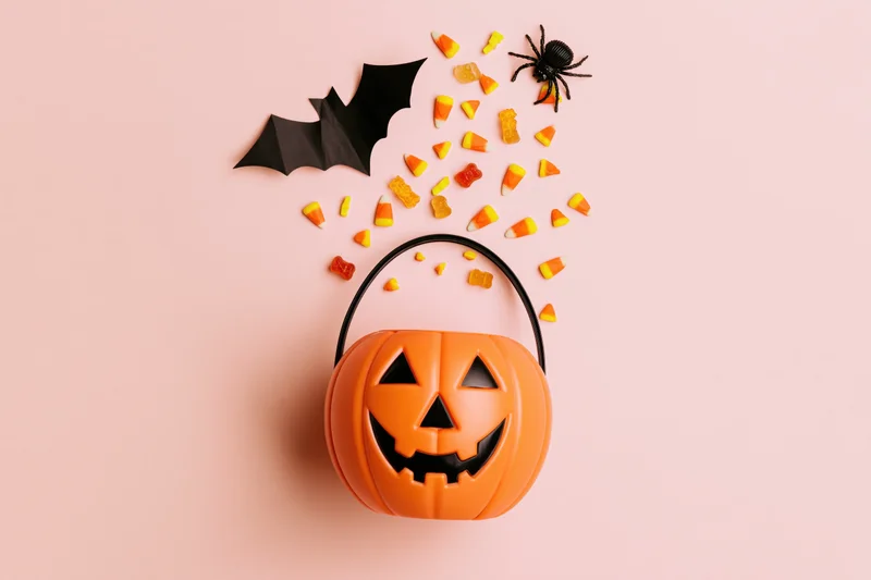 Jack-o-lantern candy bucket with scattered candy, bat, and spider on a pink background