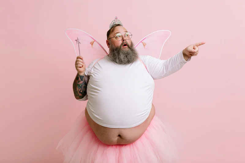 Heavyset bearded man in tooth fairy costume pointing with surprised expression against pink backdrop