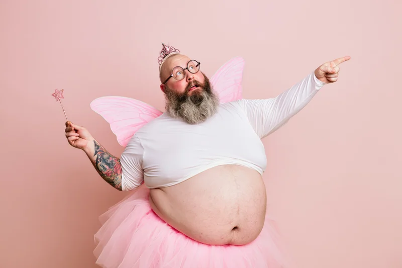 Heavyset bearded man in tooth fairy costume pointing with wand against pink background