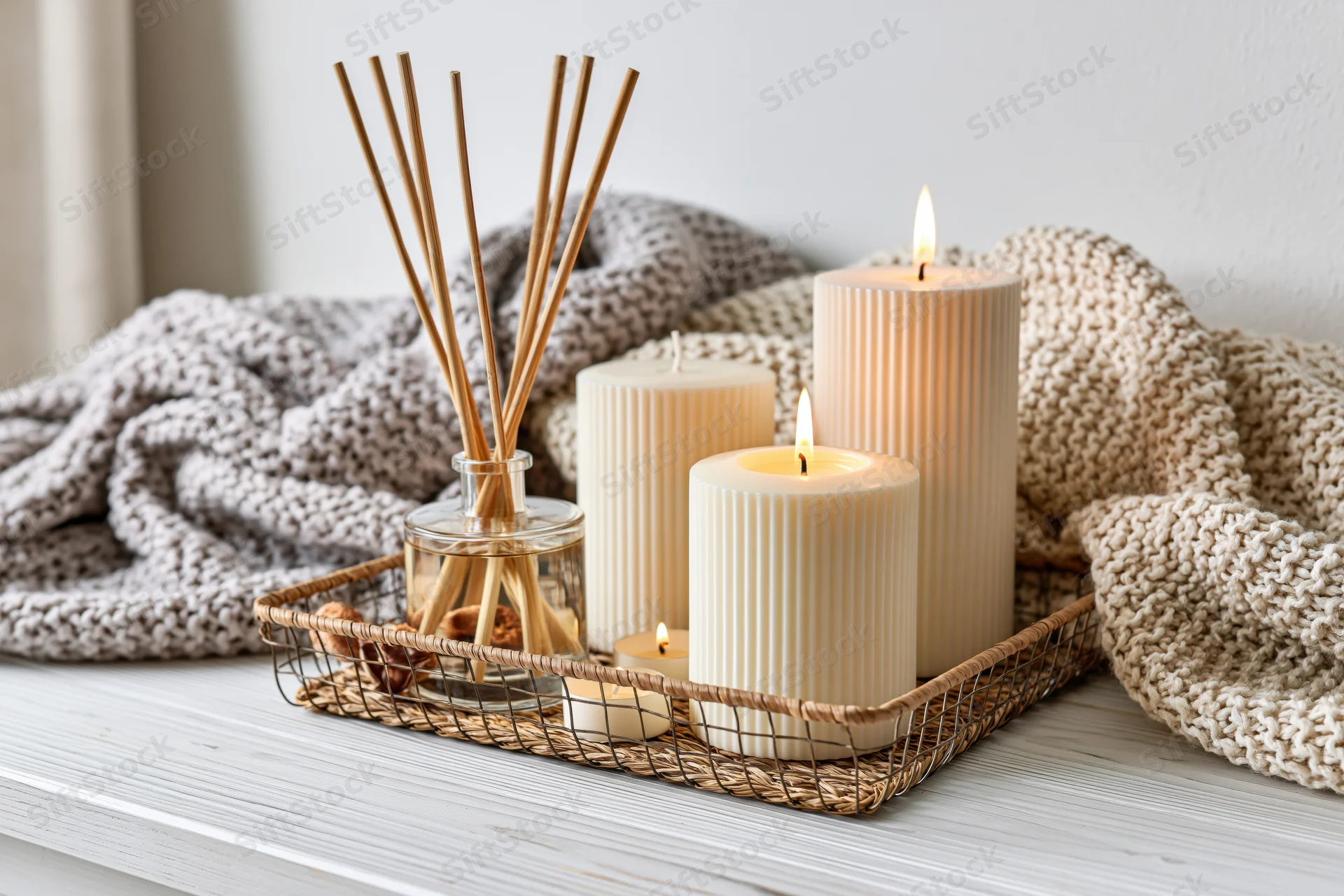 Lit candles and reed diffuser arranged in a wire basket on a white shelf with knitted blankets nearby