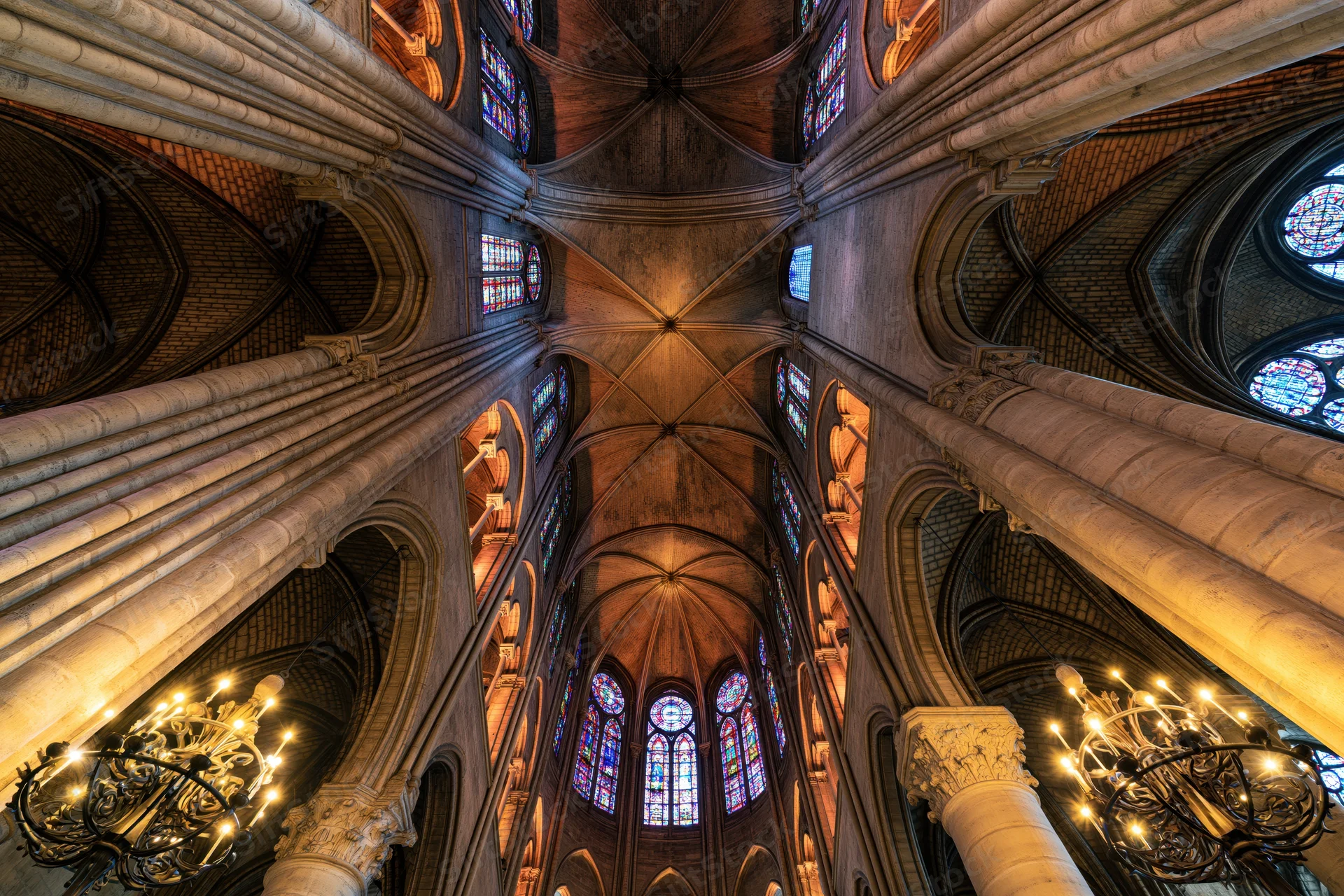 Upward view of a gothic cathedral interior with vaulted stone ceilings, stained glass windows, and ornate chandeliers