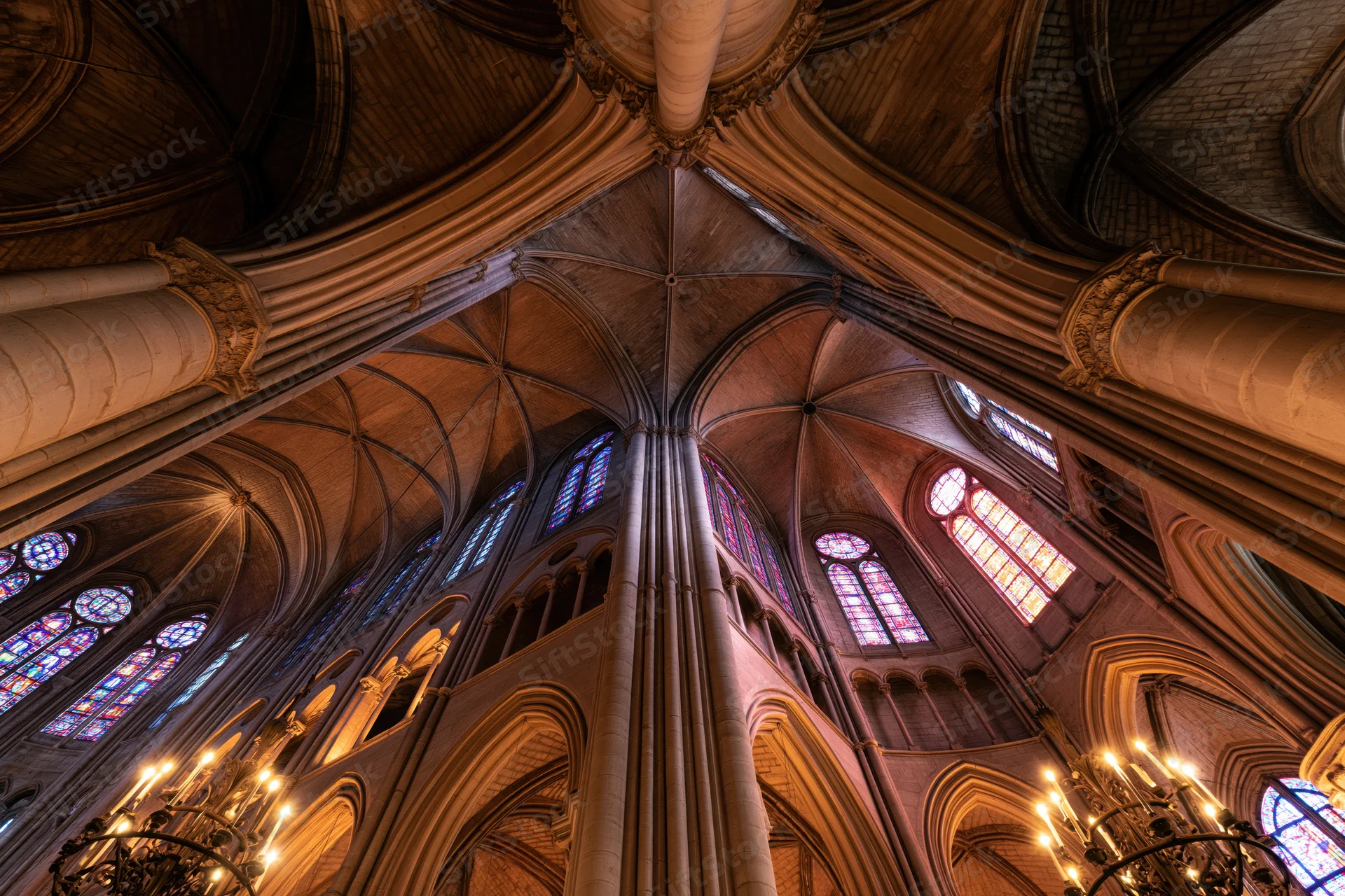 Upward view of a gothic cathedral interior with vaulted stone ceiling, stained glass windows, and chandeliers