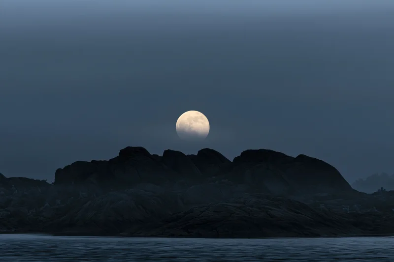 Full moon rising over rocky coastline at twilight with foggy sky and calm sea