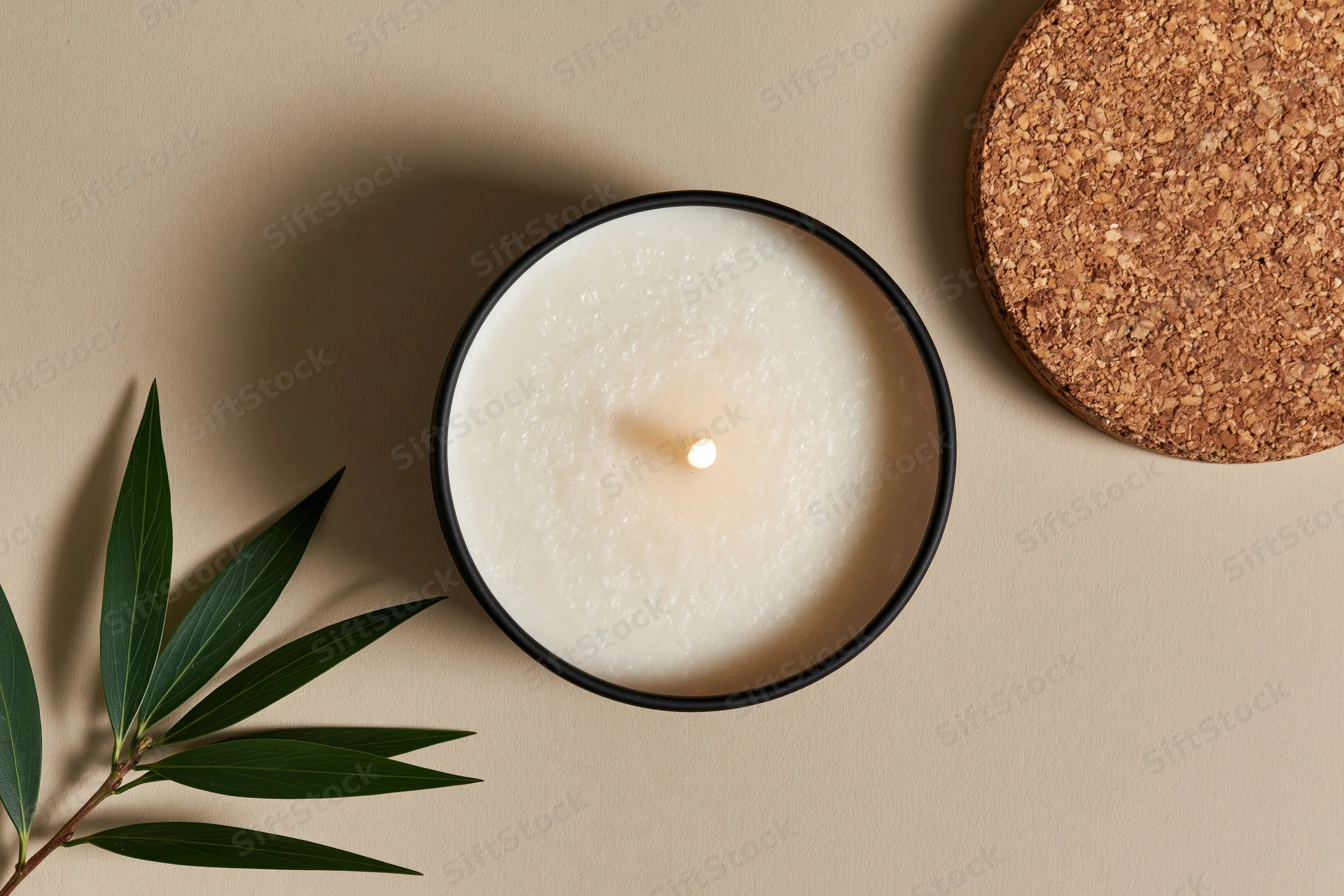 Lit soy candle in a matte black tin container with green leaves and cork coaster on beige surface