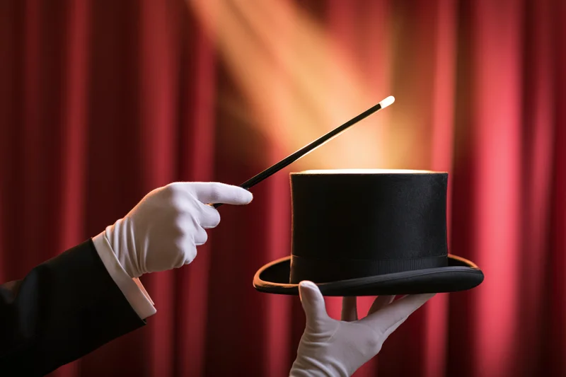 Magician's gloved hands holding a black top hat and pointing a wand against a red curtain backdrop