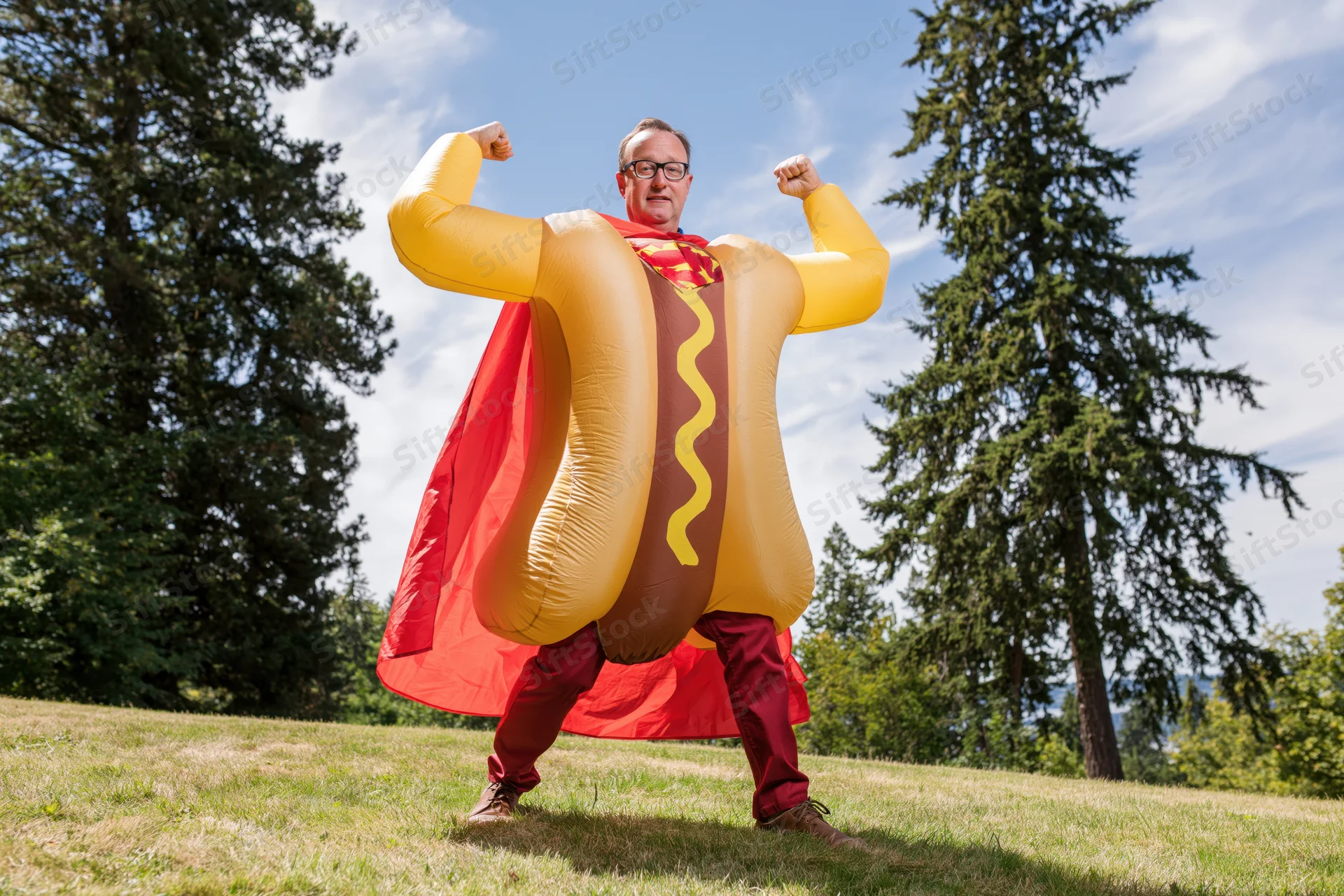 Man in inflatable hot dog superhero costume flexing arms outdoors on grassy field
