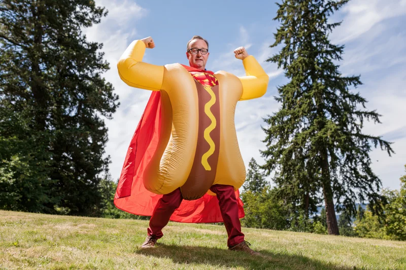 Man in inflatable hot dog superhero costume flexing arms outdoors on grassy field