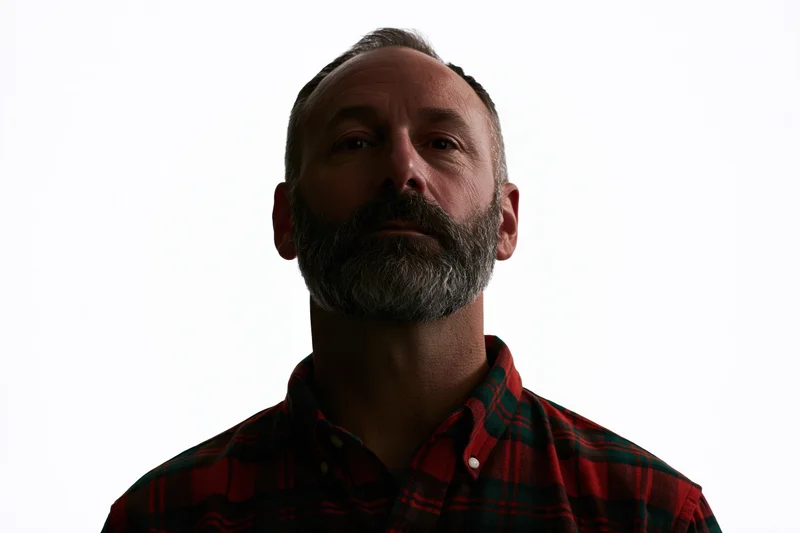 Middle-aged man with grey beard and plaid shirt standing still in studio lighting against white background