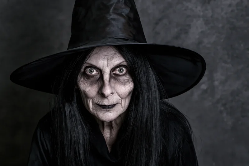 Old woman in black witch costume with wide eyes staring at camera in studio setting