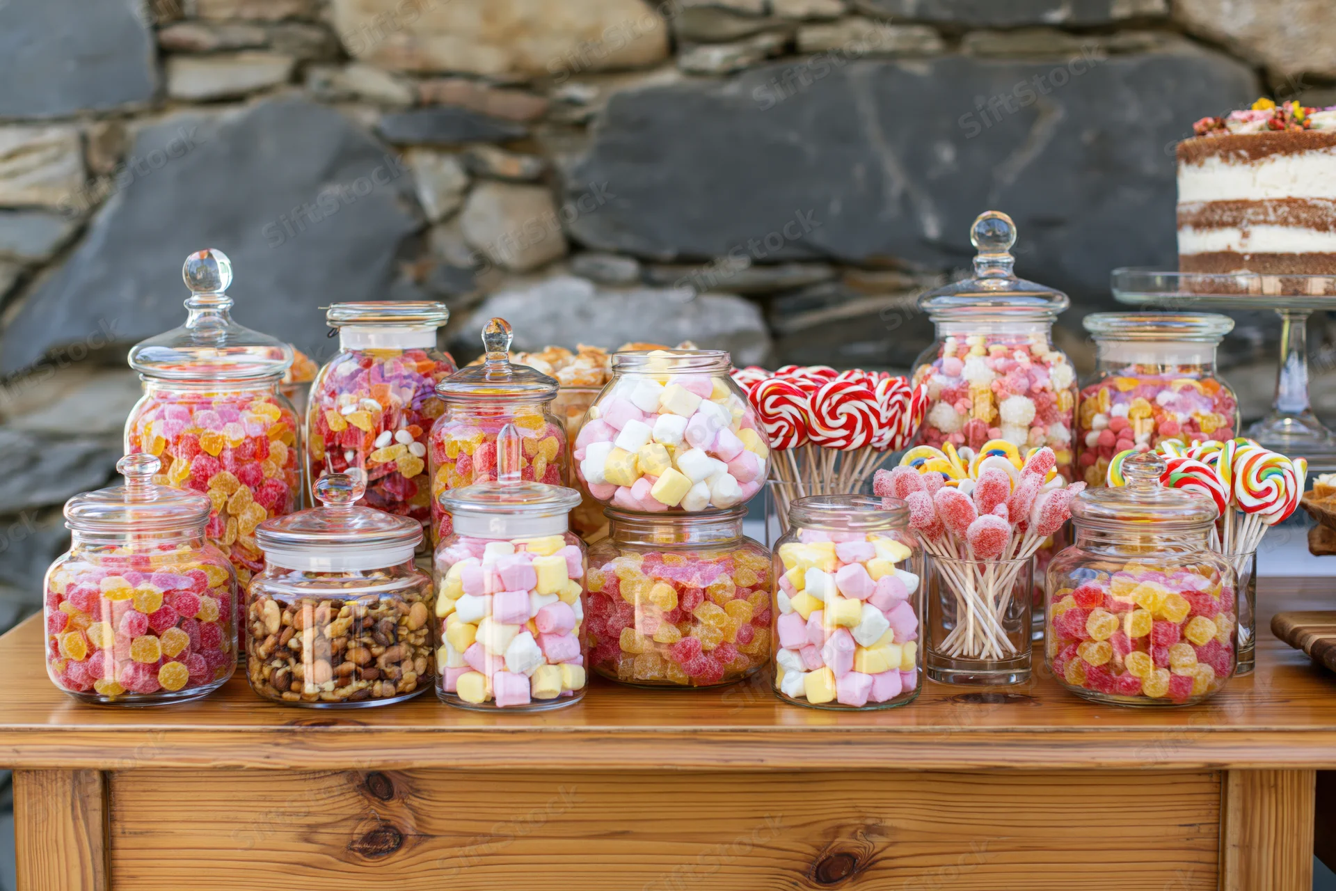 Candy jars filled with sweets arranged on a rustic wooden table outdoors near a stone wall with a tiered cake in the background
