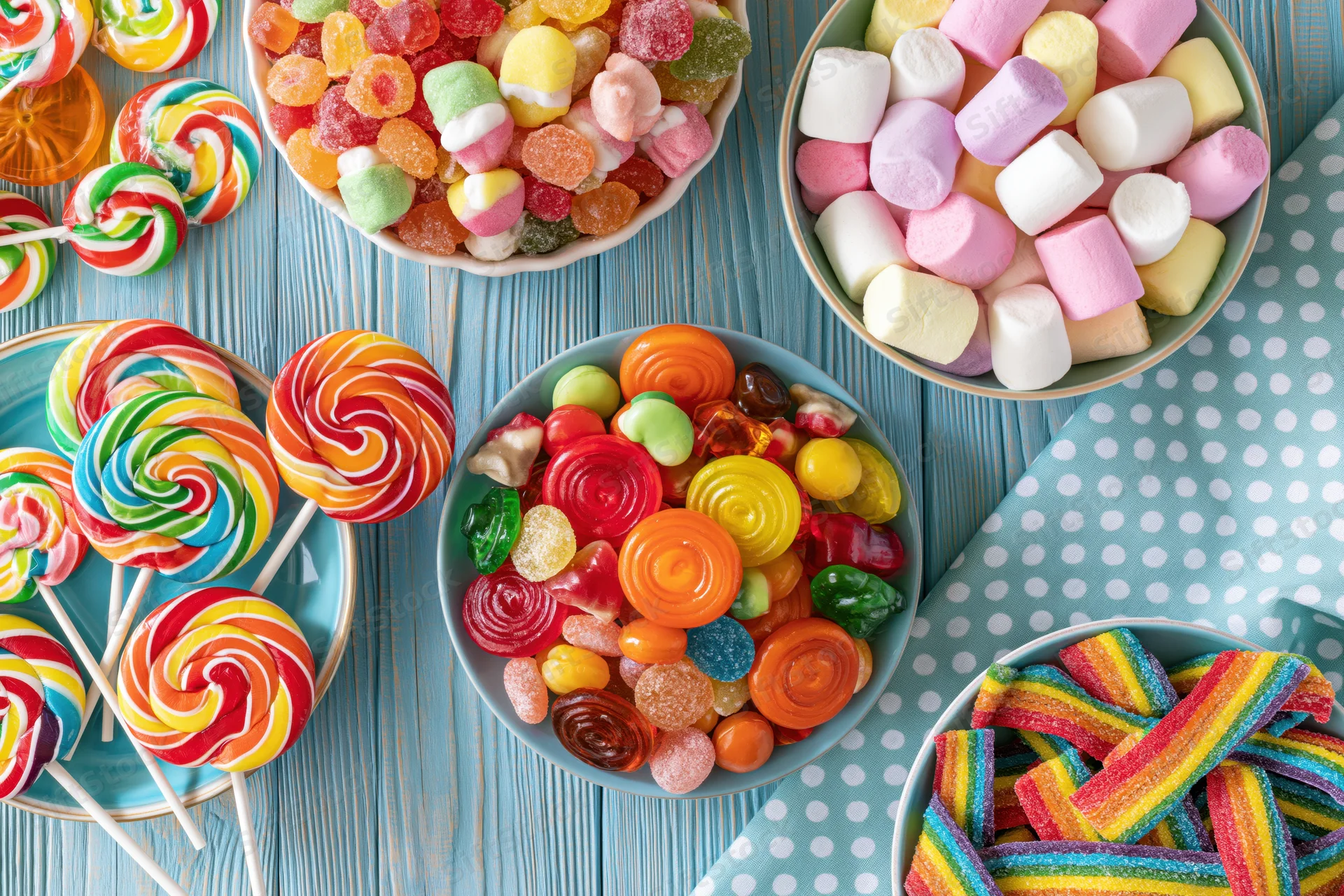 Colorful assorted candies and lollipops arranged in bowls and on plates on a blue wooden table with polka dot napkin