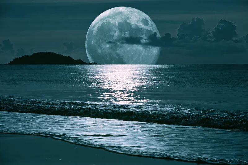 Full moon rising over calm ocean at night with reflection on water surface near shore