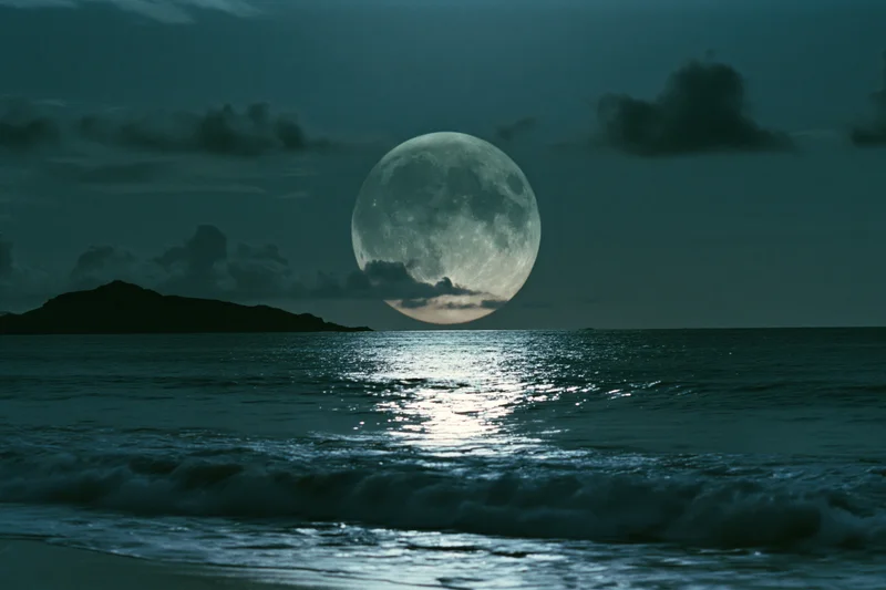 Full moon rising over calm ocean at night with reflection on water and distant island silhouette