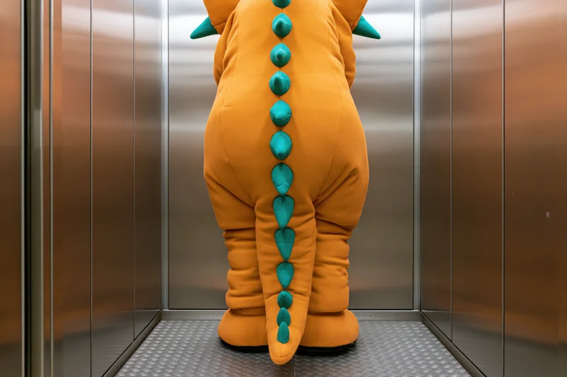 Back view of a person in an orange and green dinosaur mascot costume standing in a stainless steel elevator