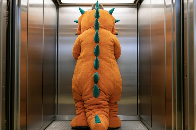 Person in orange and green dinosaur mascot costume standing in elevator facing away