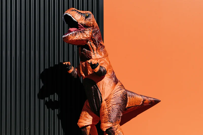 Person in inflatable t-rex costume standing against black corrugated wall and orange background