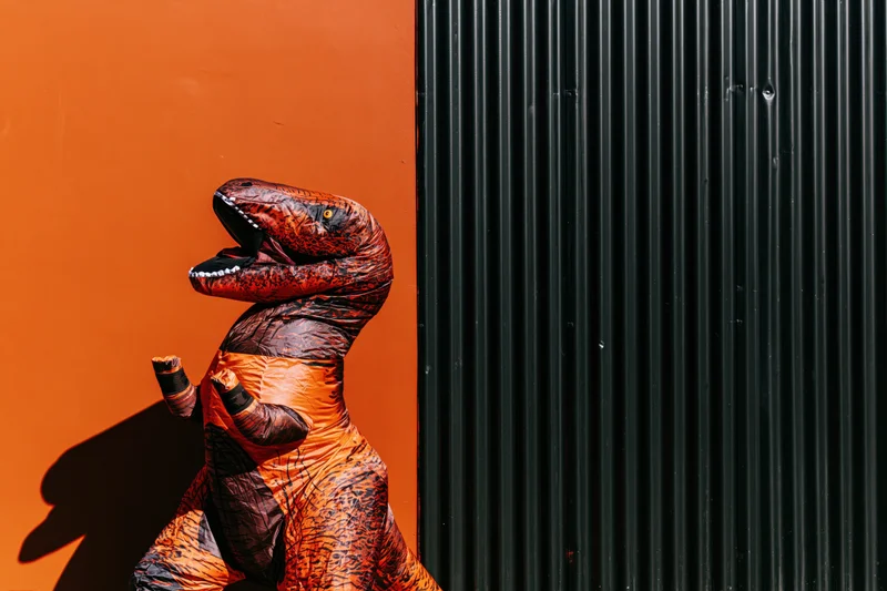 Person in inflatable t-rex costume standing against orange wall and black corrugated metal panel