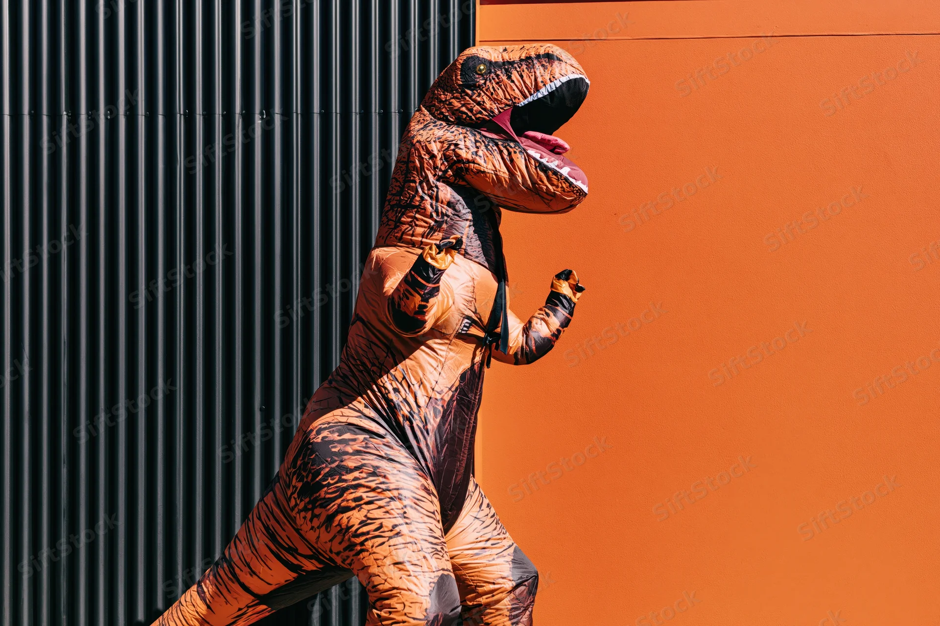 Person in inflatable t-rex costume running against orange and black wall outdoors