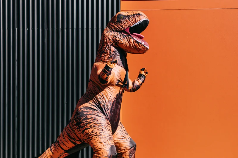 Person in inflatable t-rex costume running against orange and black wall outdoors