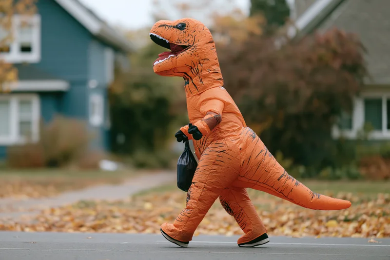 Person in inflatable t-rex costume walking on suburban street with candy bag during halloween