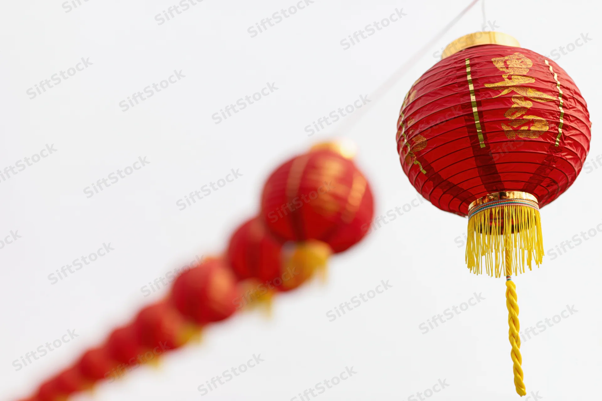 Row of red paper lanterns with golden characters and tassels hanging on a string against a bright sky