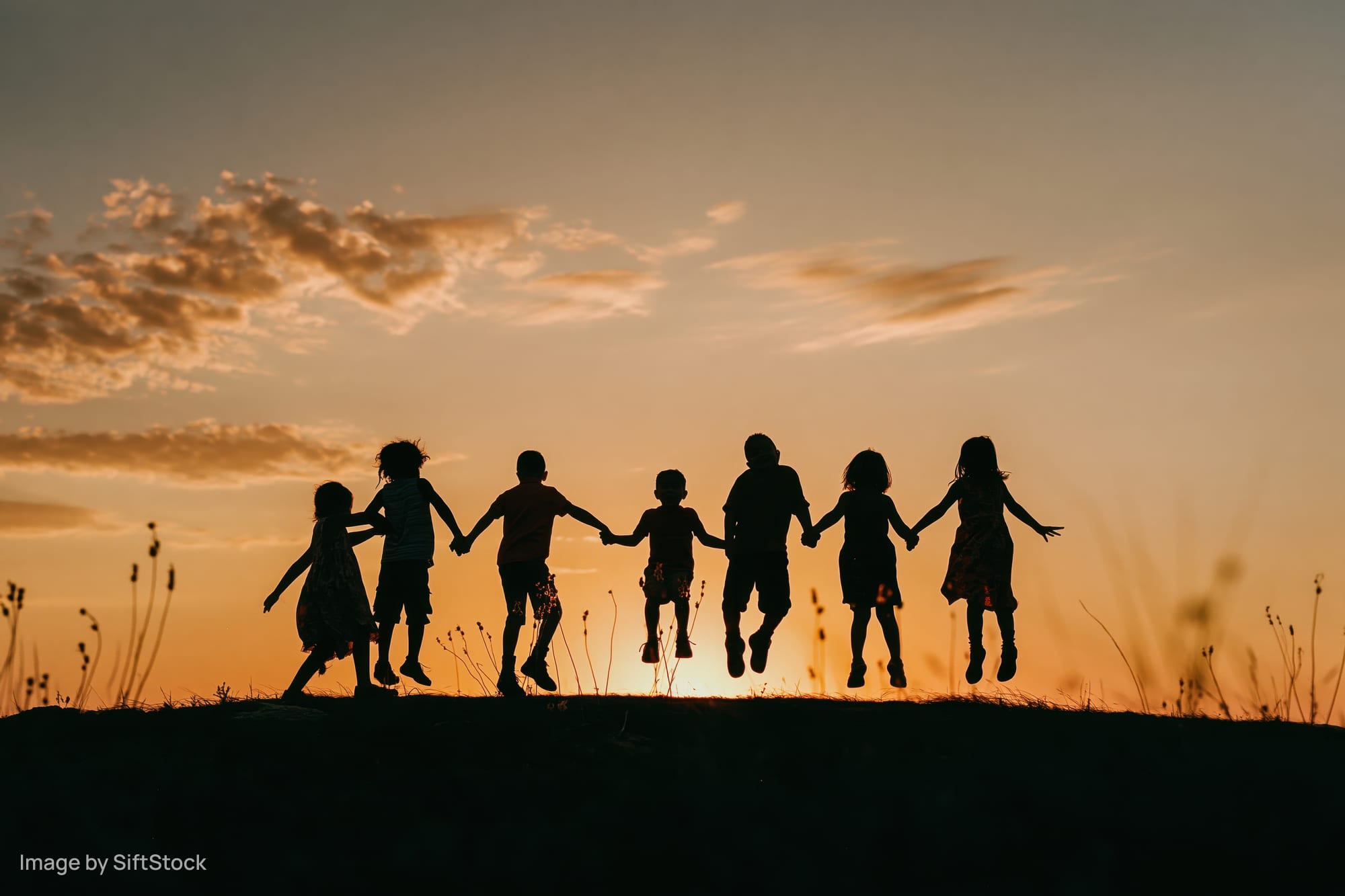 Silhouetted children holding hands and jumping on a hill at sunset