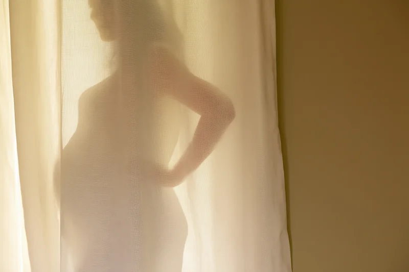 Pregnant woman’s silhouette cast on sheer linen curtain by soft window light