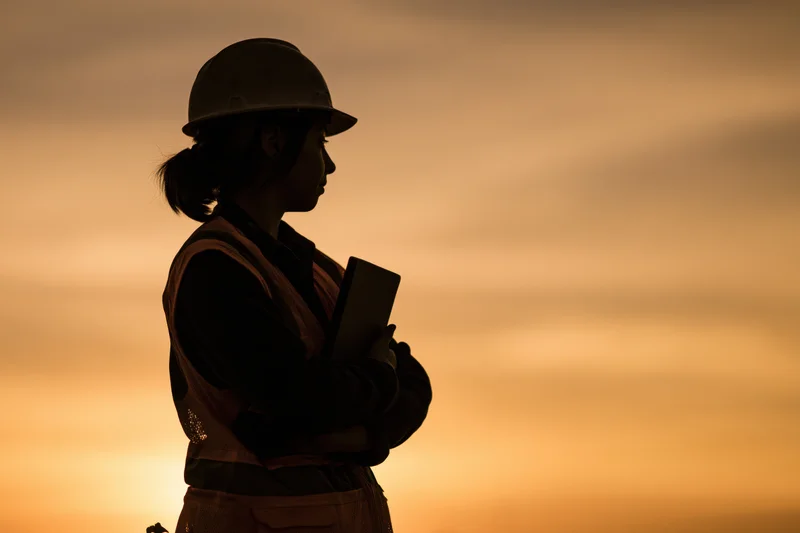 Silhouette of a female construction engineer holding a tablet outdoors at sunset