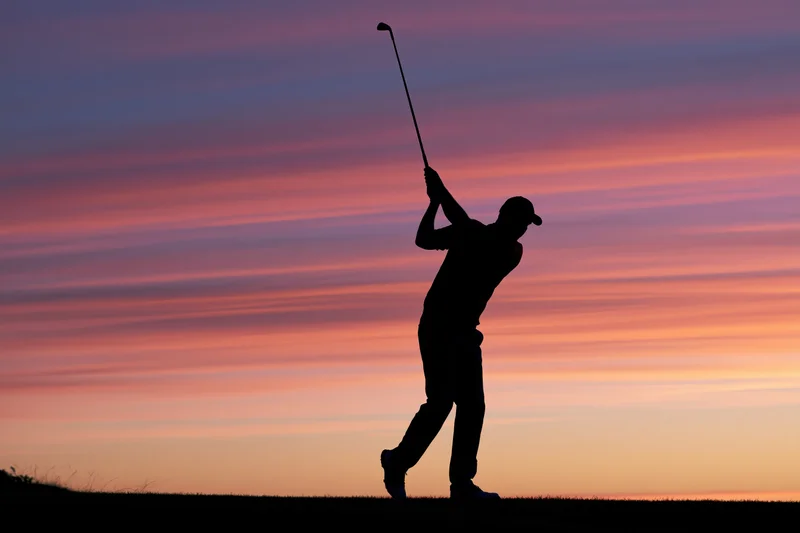 Silhouette of a golfer completing a full follow-through swing at sunset on a grassy field