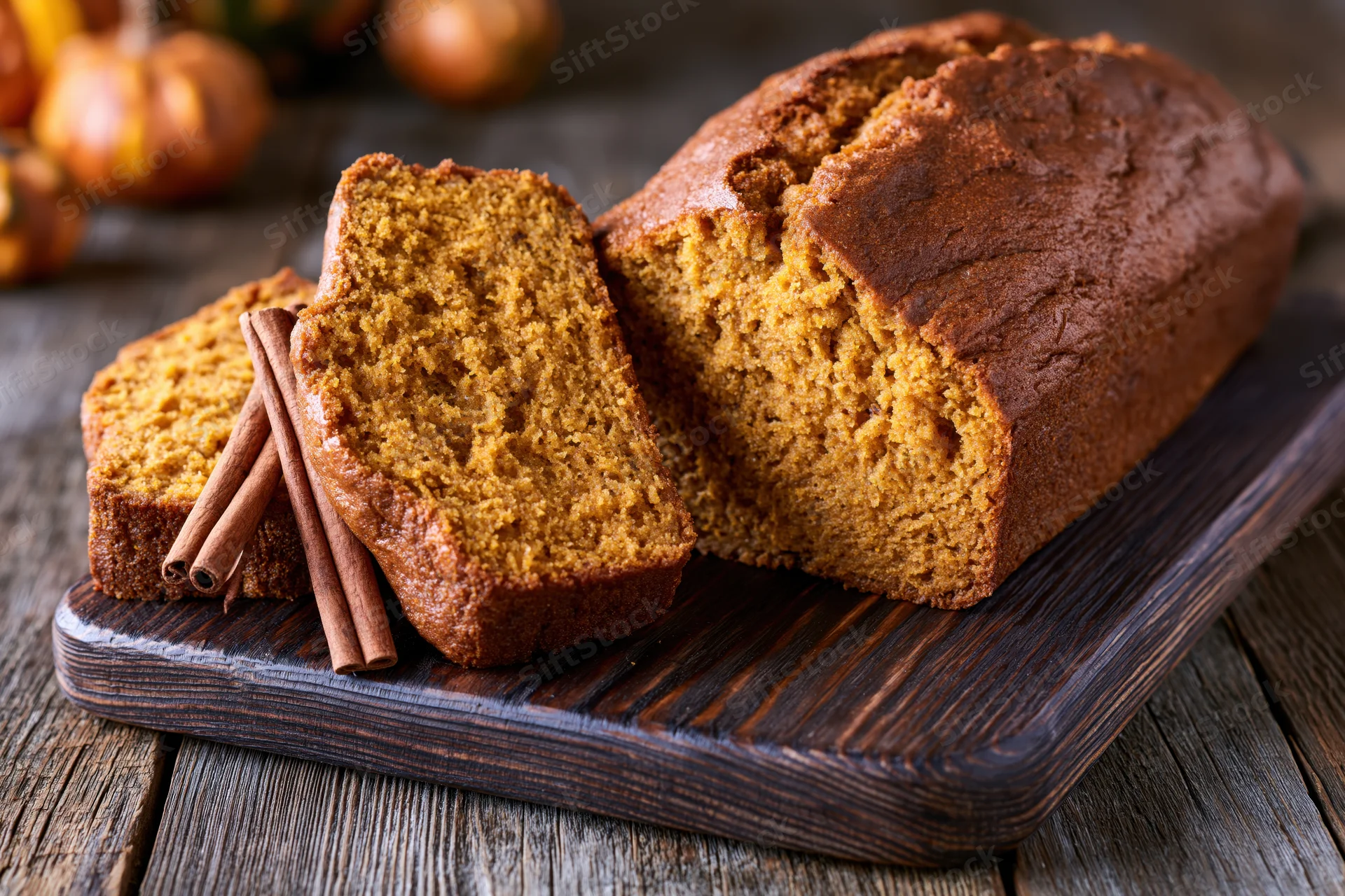 Sliced pumpkin loaf cake on dark wooden cutting board with cinnamon sticks and pumpkins in background