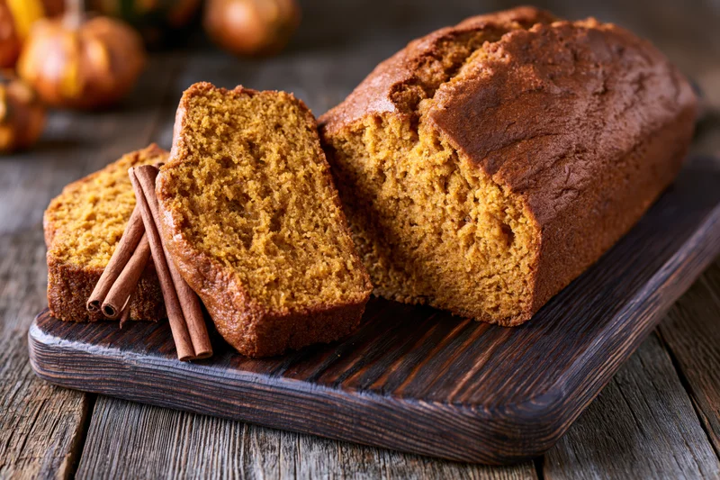 Sliced pumpkin loaf cake on dark wooden cutting board with cinnamon sticks and pumpkins in background