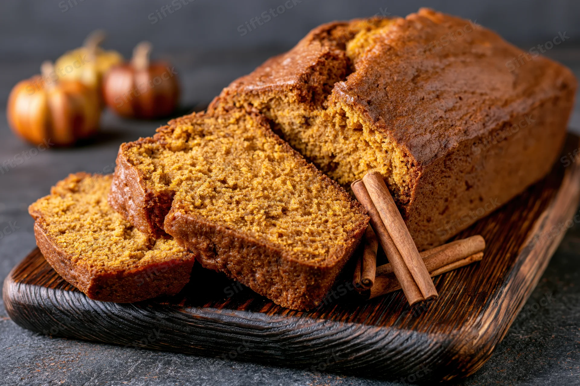 Sliced pumpkin loaf cake on dark wooden cutting board with cinnamon sticks and small pumpkins in background