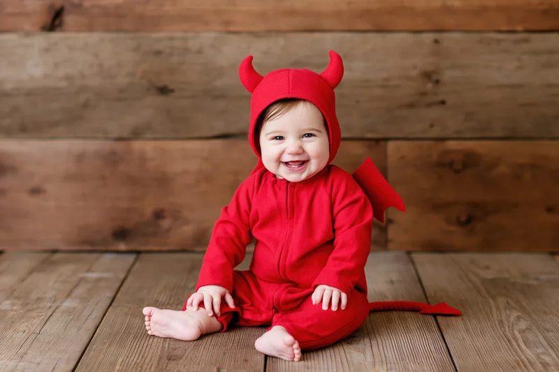 Smiling baby in red devil costume sitting on wooden floor