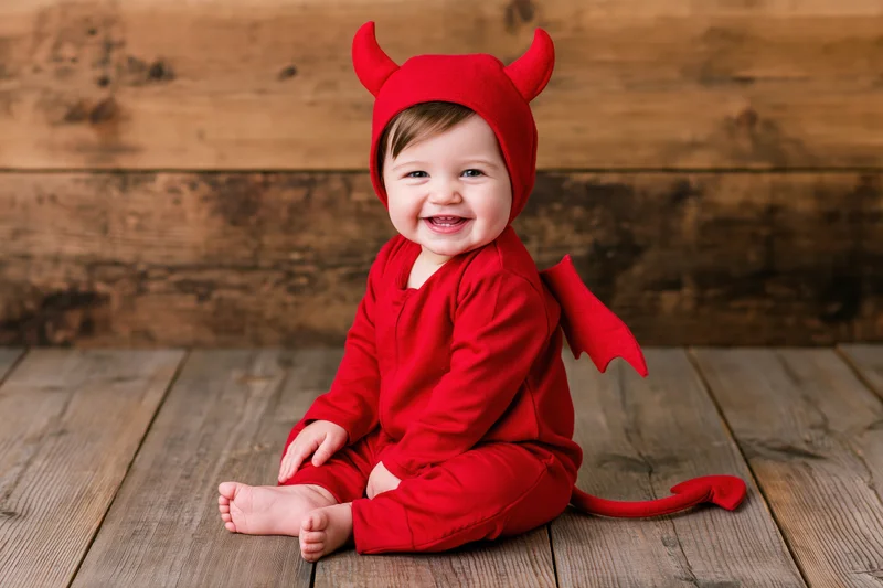 Smiling baby in red devil costume sitting on wooden floor