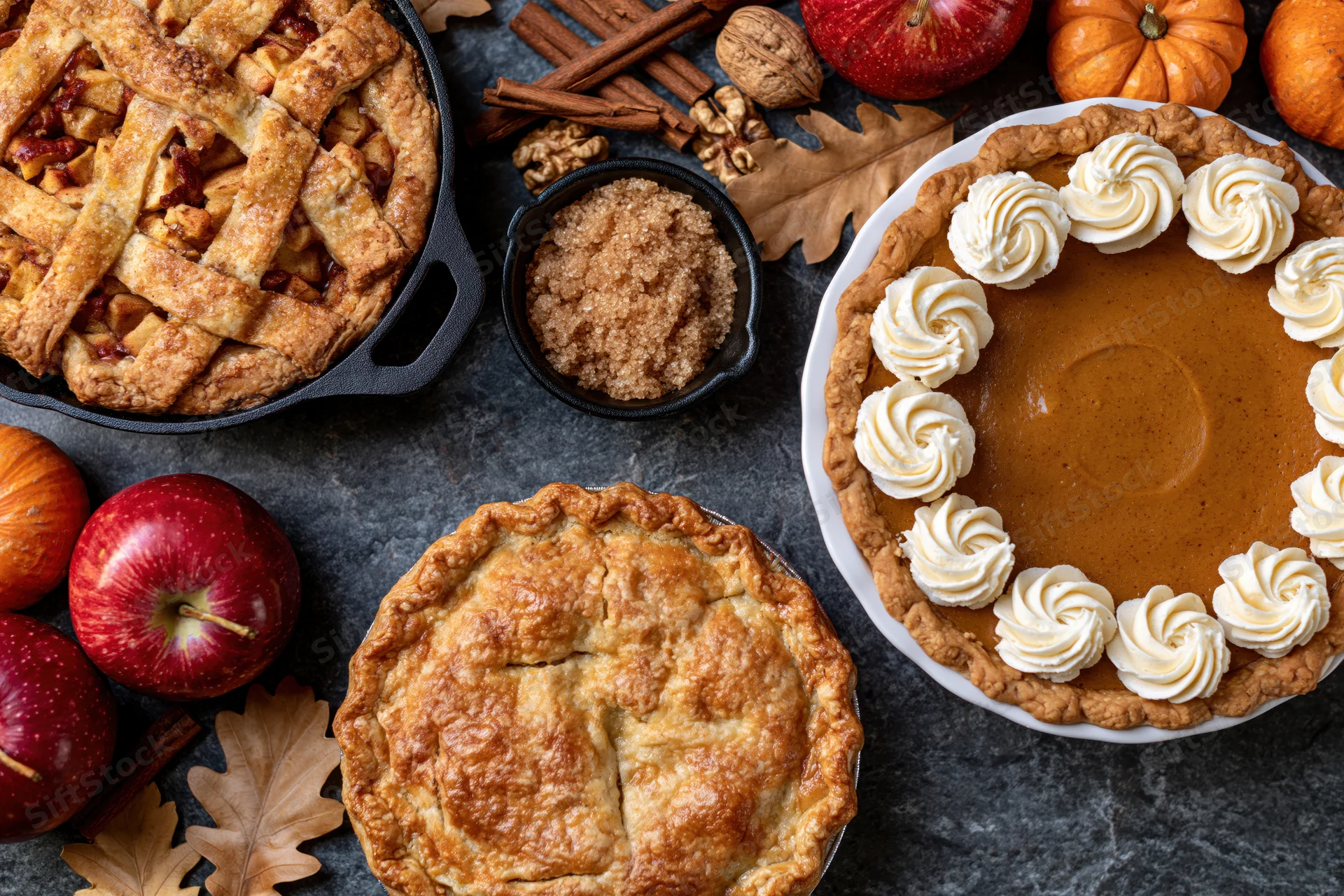 Three autumn pies arranged on dark stone surface with apples, pumpkins, and spices