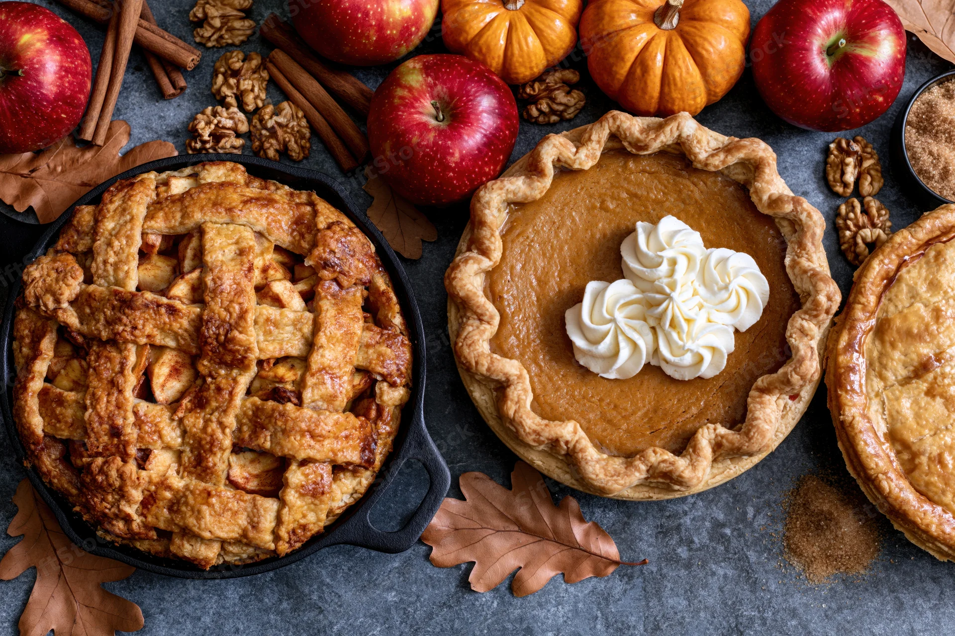 Three autumn pies arranged on a dark stone surface with apples, pumpkins, and fall leaves