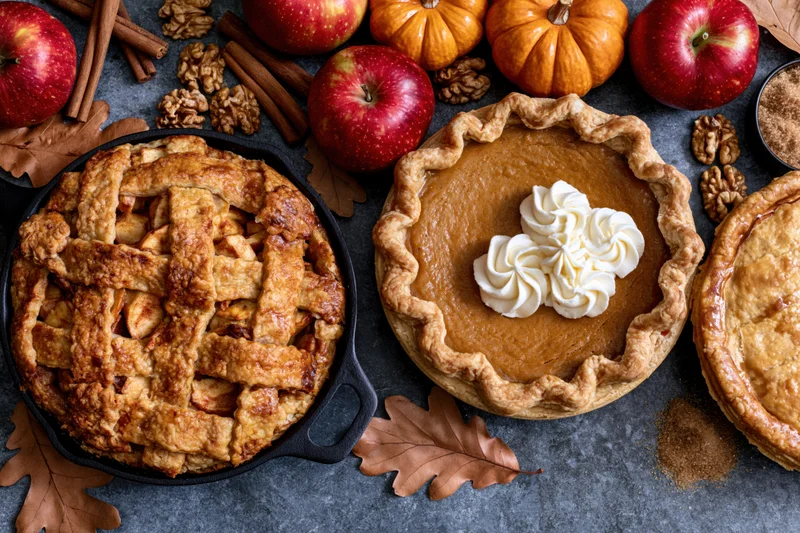 Three autumn pies arranged on a dark stone surface with apples, pumpkins, and fall leaves