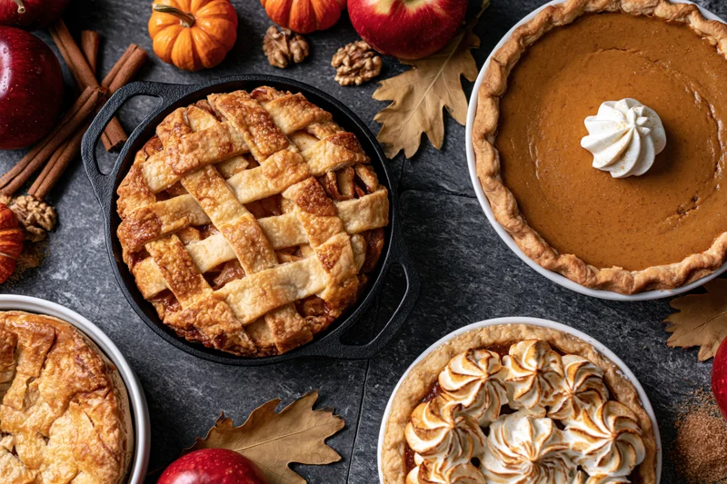 Three autumn pies arranged on dark stone surface with apples, pumpkins, and cinnamon sticks