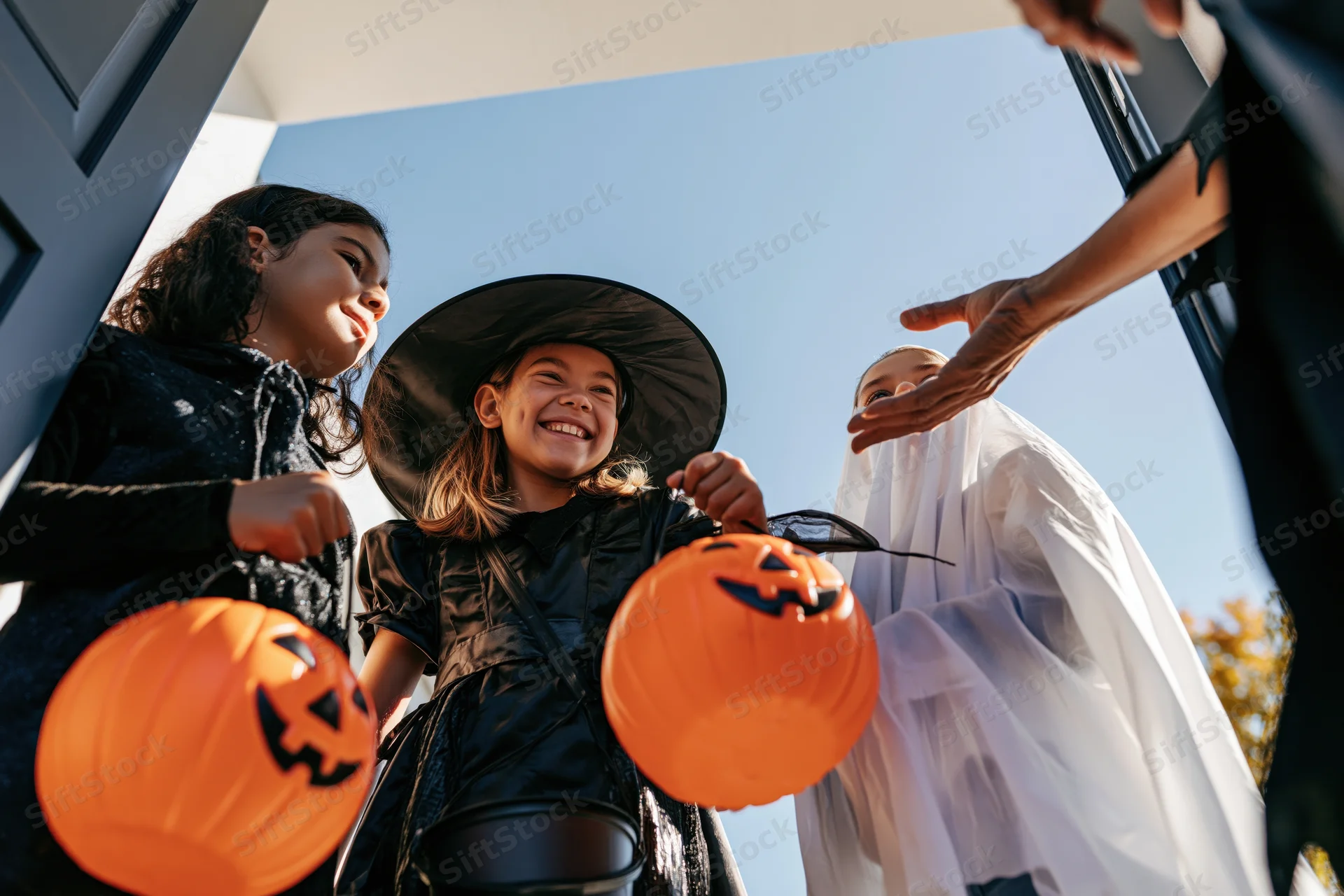 Three children in halloween costumes trick-or-treating at a house door, smiling and holding pumpkin buckets