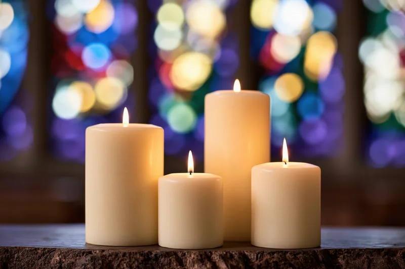 Four ivory pillar candles burning on a stone surface in front of stained glass windows in a church interior