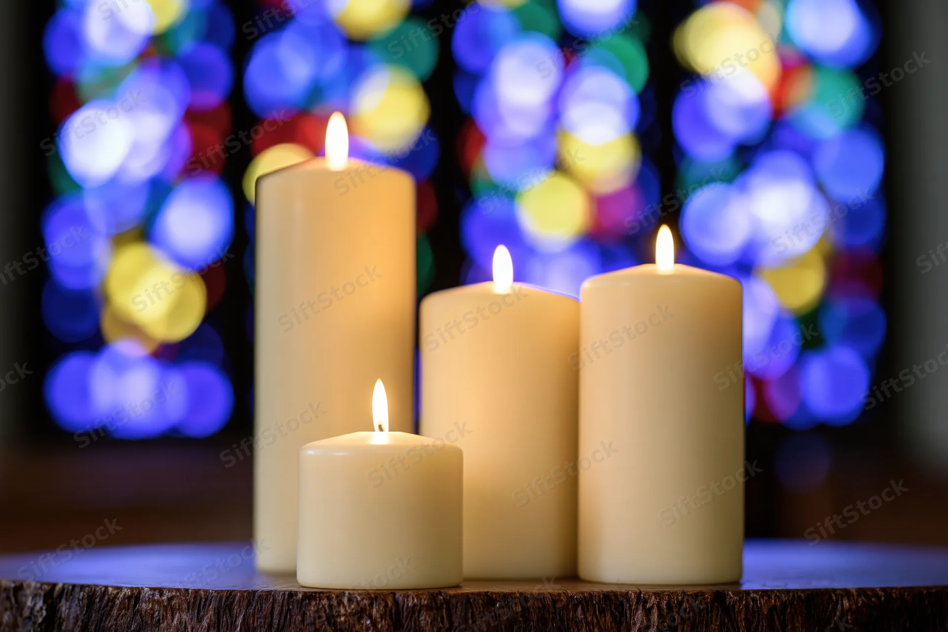 Four ivory candles burning on a wooden surface with stained glass bokeh in the background