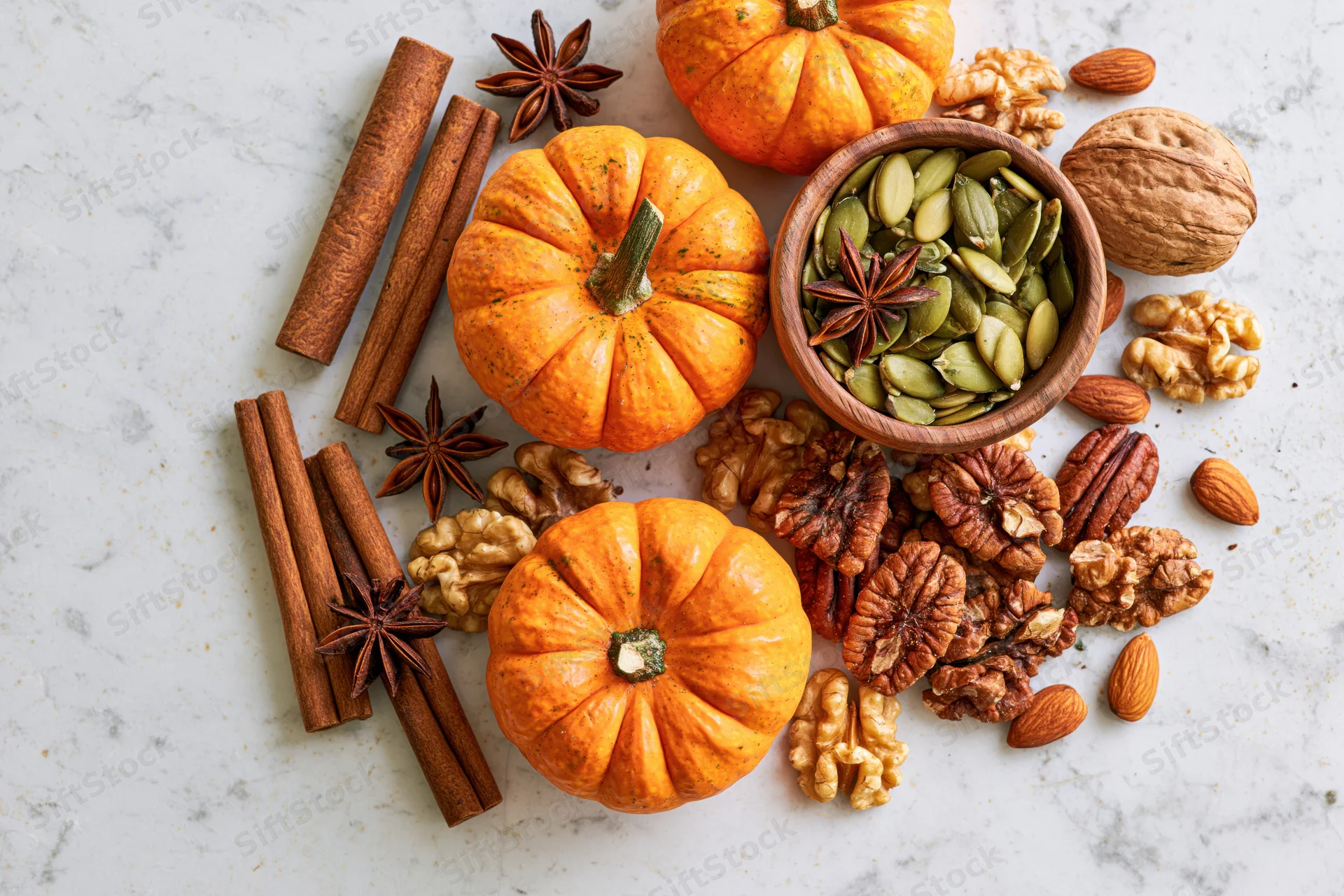 Small orange pumpkins with cinnamon sticks, nuts and seeds arranged on marble surface
