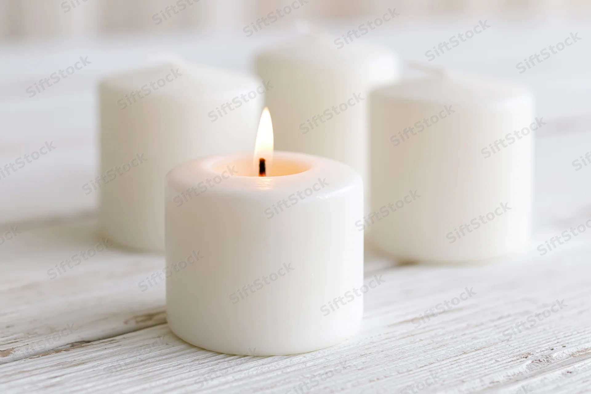 Lit white pillar candle on a white painted wooden table with other candles in soft focus