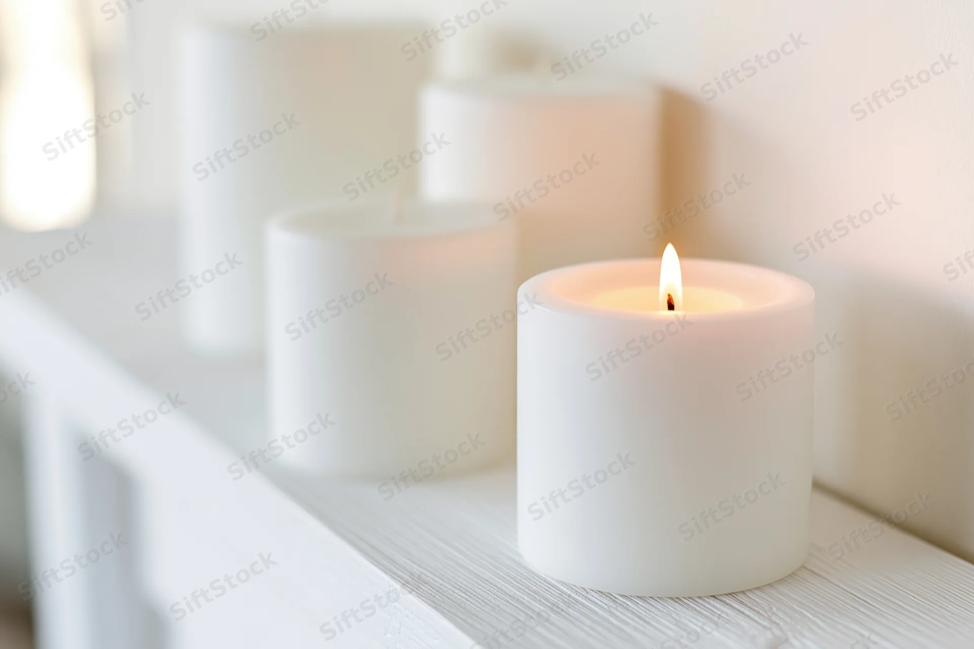 Lit white pillar candle on a white wooden shelf with other candles in soft focus