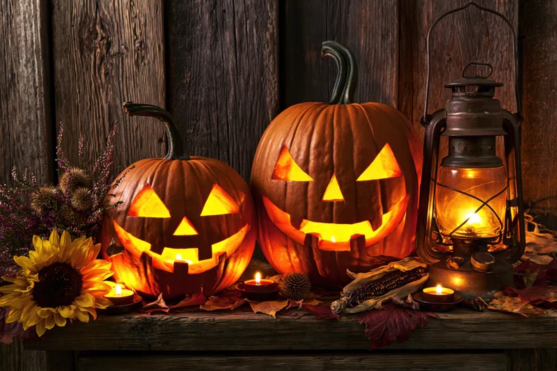 Two carved jack-o-lanterns glowing with candles on a rustic wooden table surrounded by autumn decorations