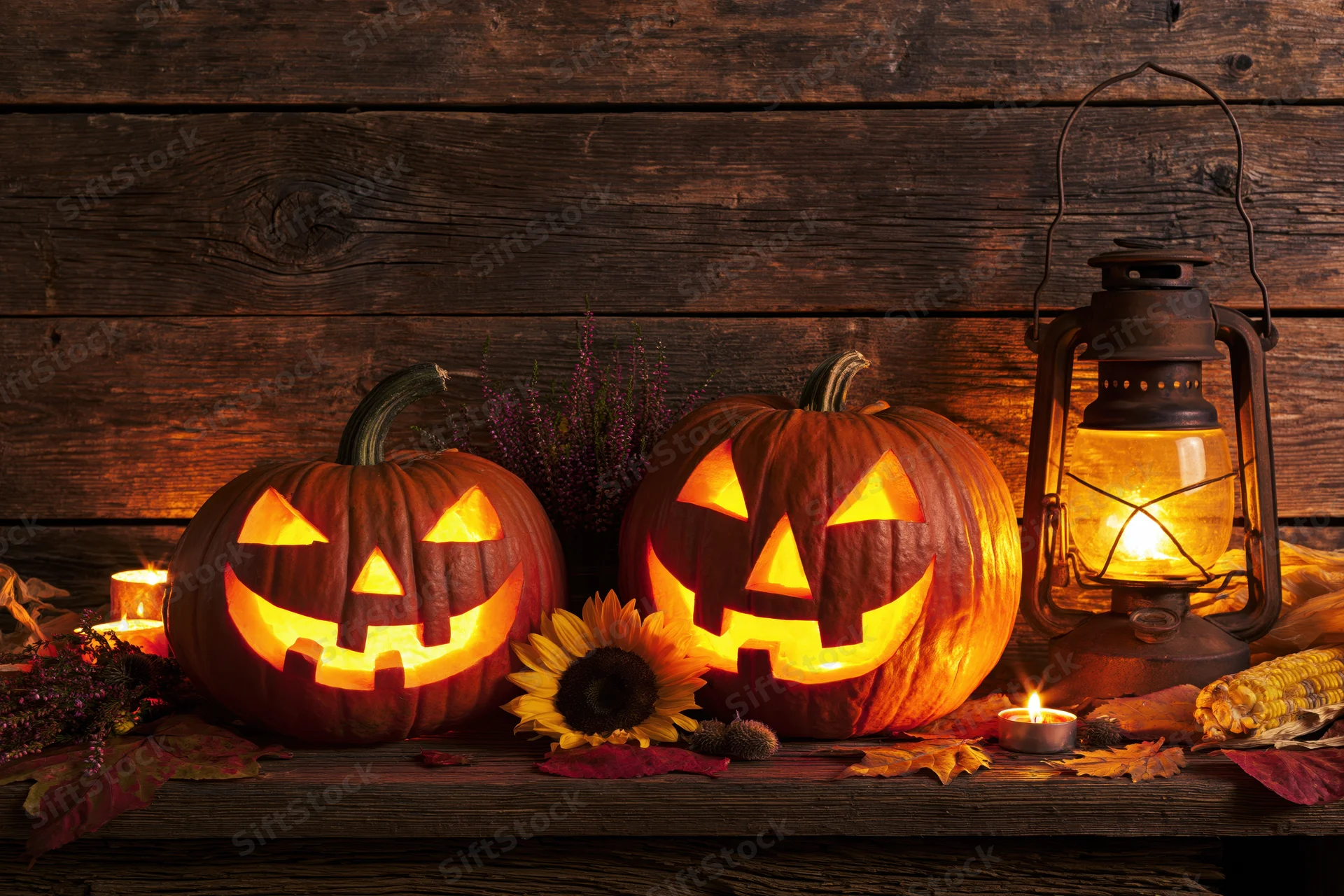 Two carved jack-o-lanterns glowing on a wooden table with candles, lantern, and autumn decorations
