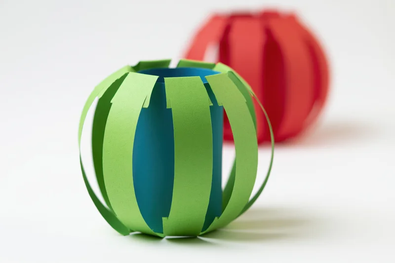 Two handmade paper lanterns in green and red on a white surface