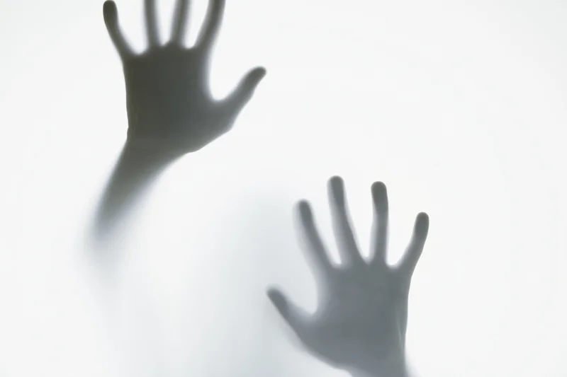 Two hands pressing against frosted glass in a blurred silhouette, creating a shadowy reach through translucent surface