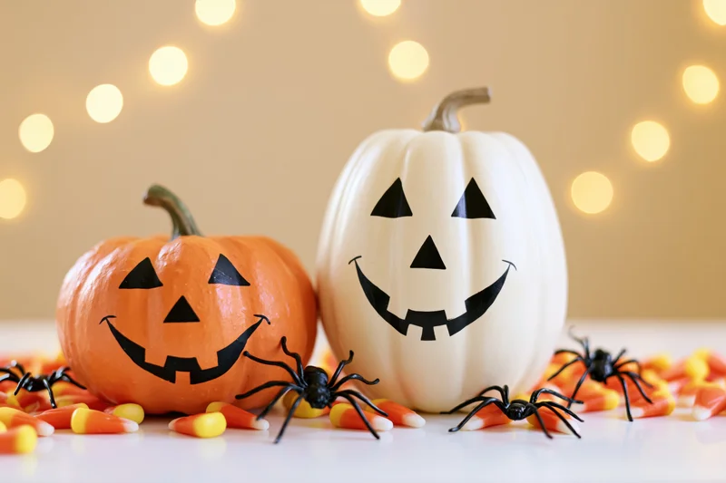 Two painted halloween pumpkins with jack-o-lantern faces on a table surrounded by candy corn and plastic spiders with warm bokeh lights in the background