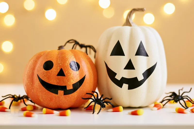 Two painted halloween pumpkins with jack-o-lantern faces on a table surrounded by candy corn and plastic spiders with warm bokeh lights in the background